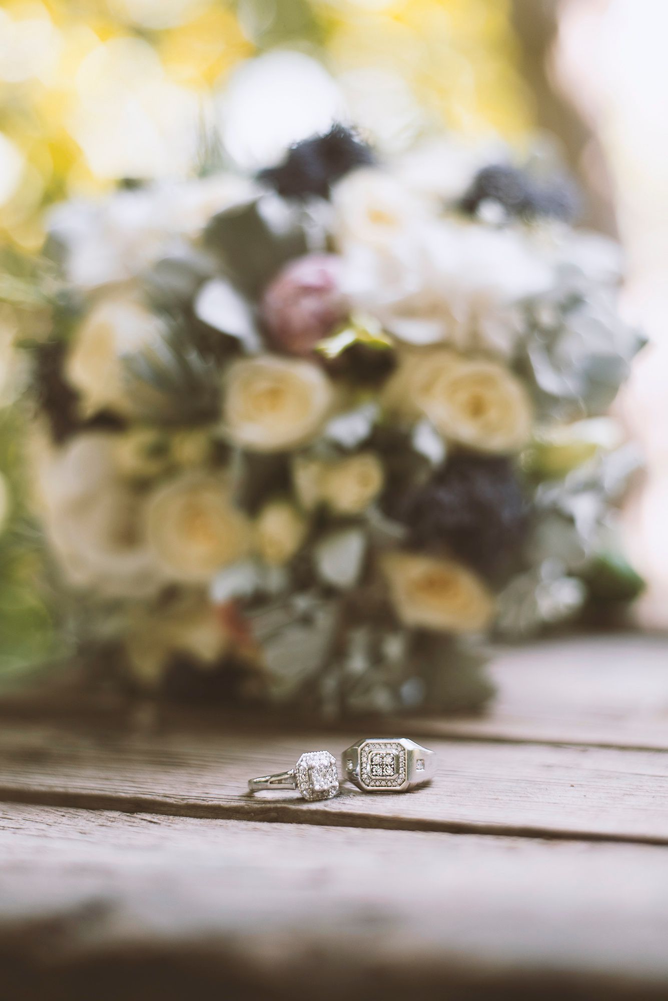 Wedding rings sit on a wooden surface with soft focus white and yellow flowers in the background.