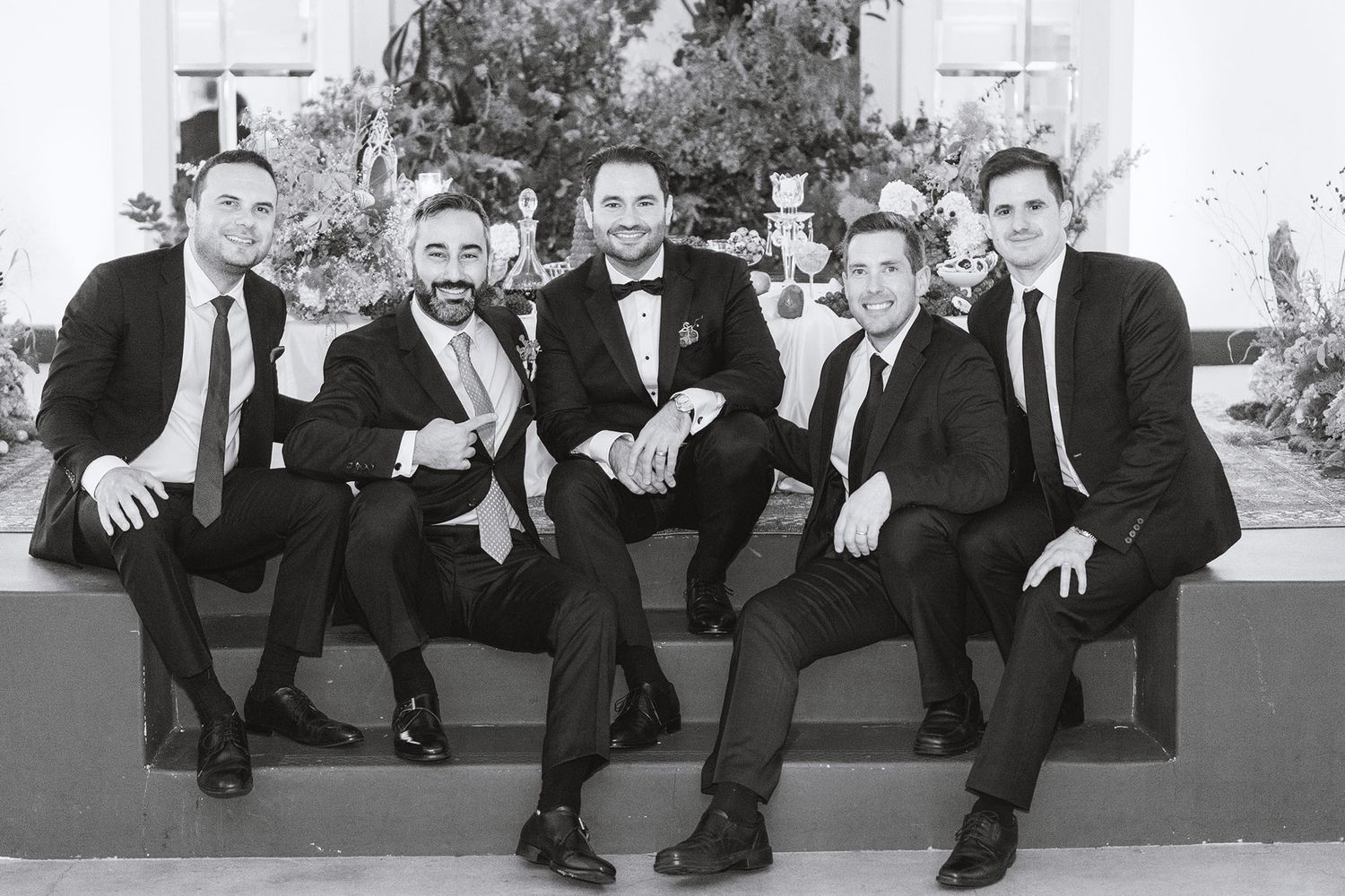 A black and white photograph shows five people in formal suits sitting casually on a ledge outdoors.
