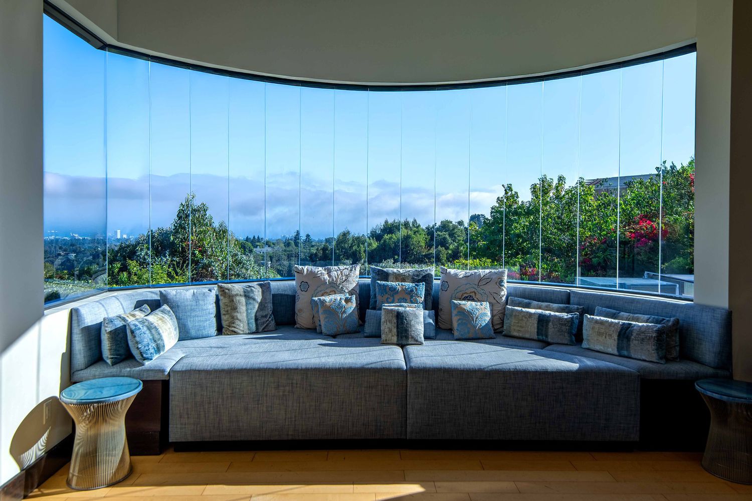 Curved window seating area with gray cushions and panoramic ocean view overlooking green hillside landscape.