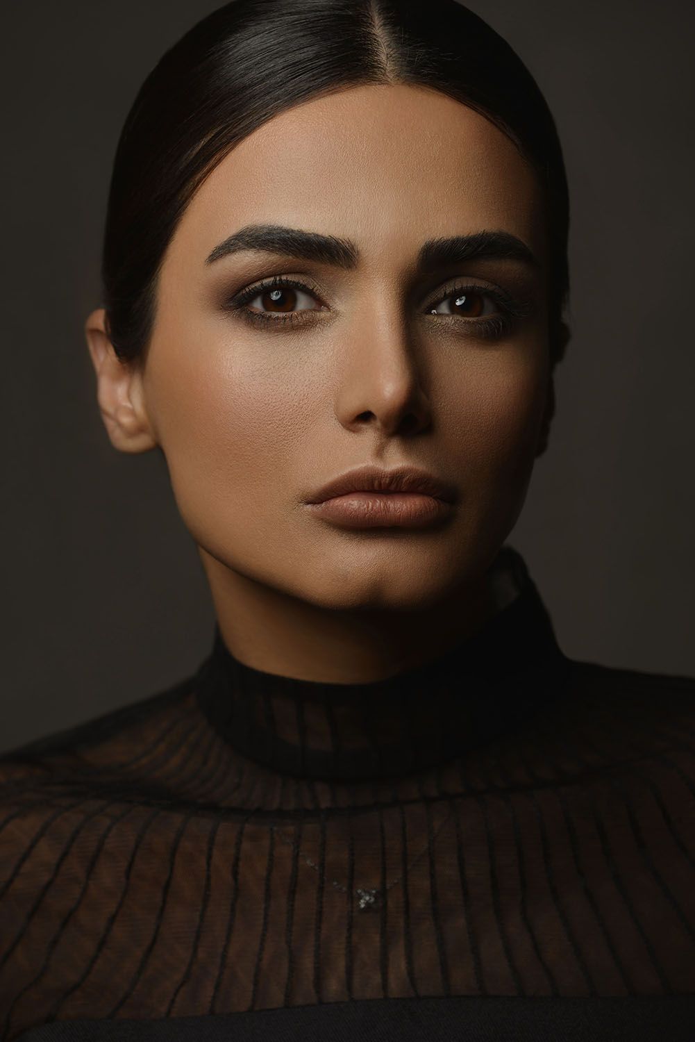 Close-up portrait of a young woman with bold brows, smoky eye makeup, and nude lips in a dark turtleneck top.