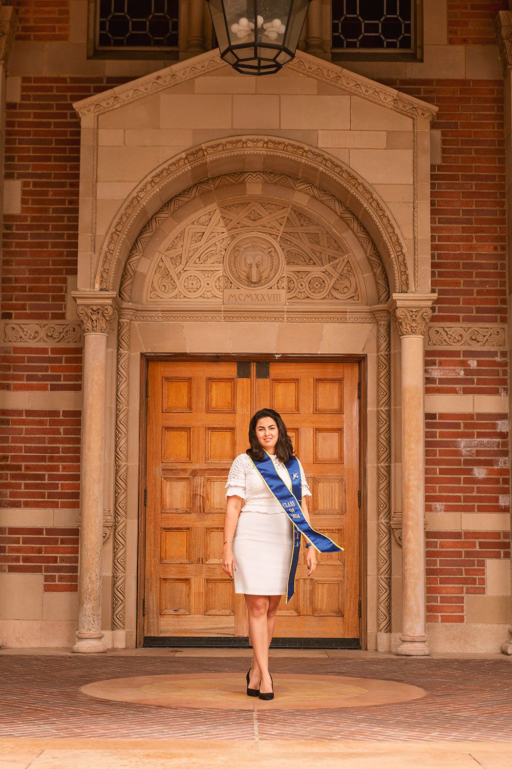 Woman in white dress with blue graduation sash stands before ornate wooden doors of a brick university building.