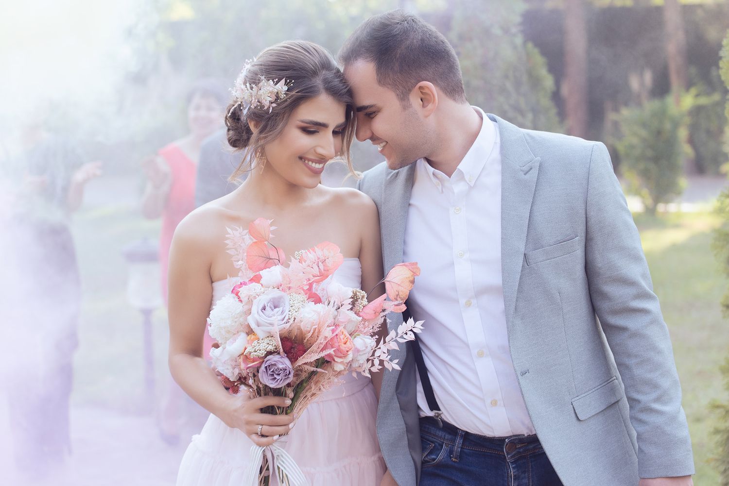 A couple in wedding attire share intimate moments in a garden setting with soft pink smoke effects in the background.