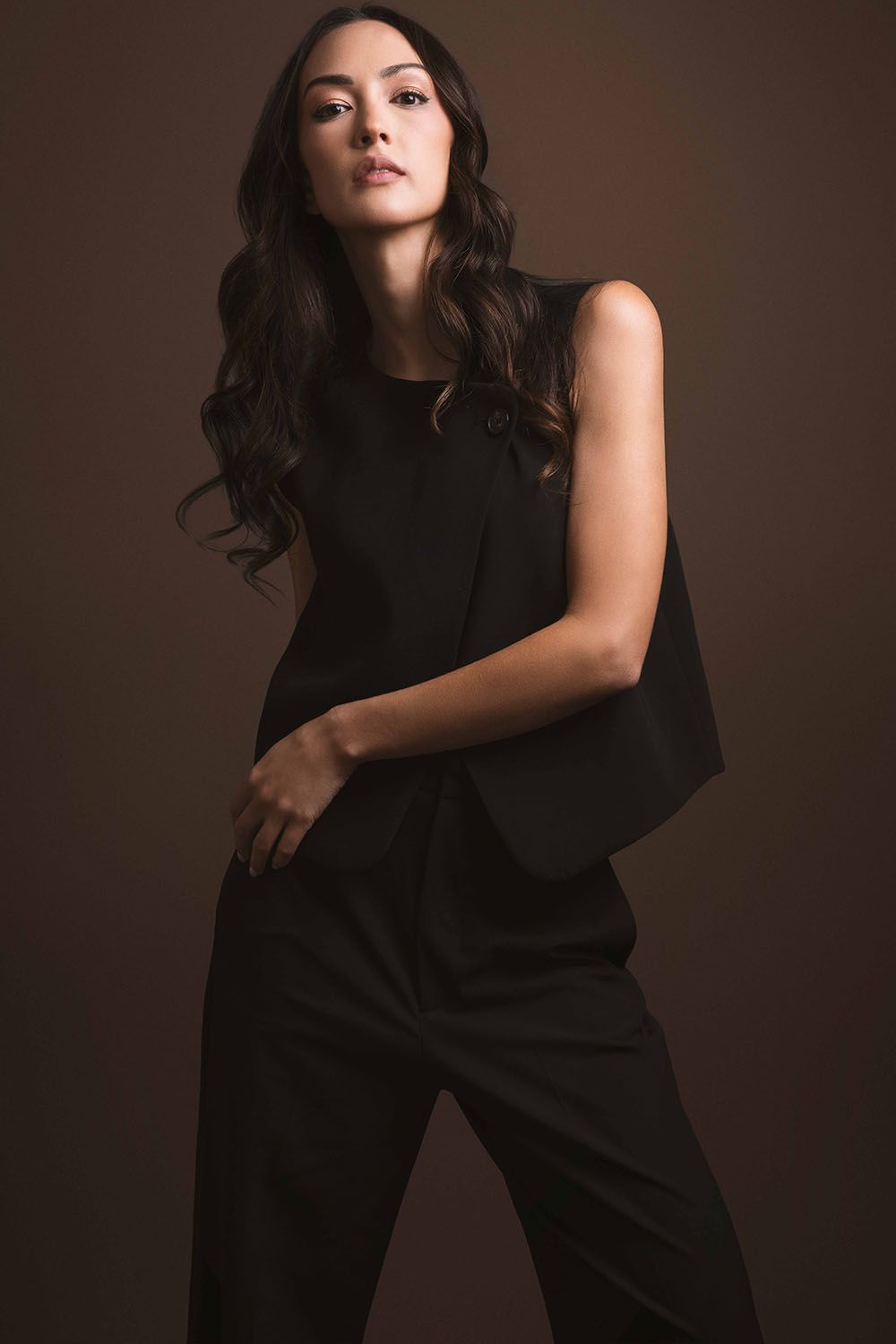Woman in sleek black sleeveless jumpsuit with cape detail, posing against a warm brown studio background.