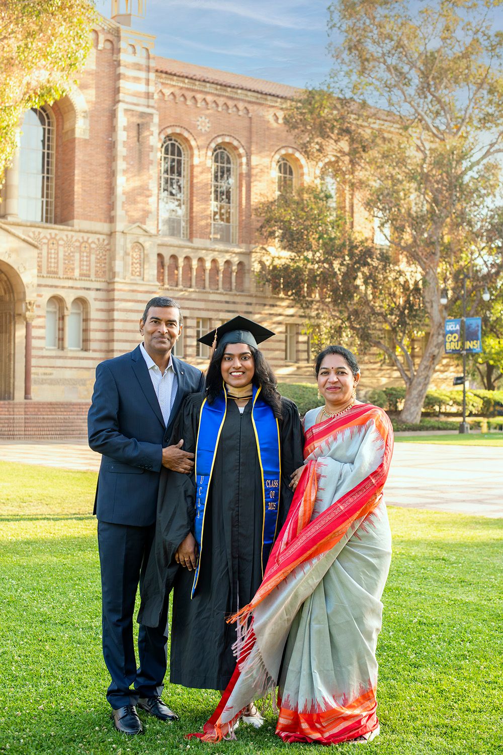 A graduate in cap and gown poses with proud parents on a sunny campus lawn in front of a brick university building.