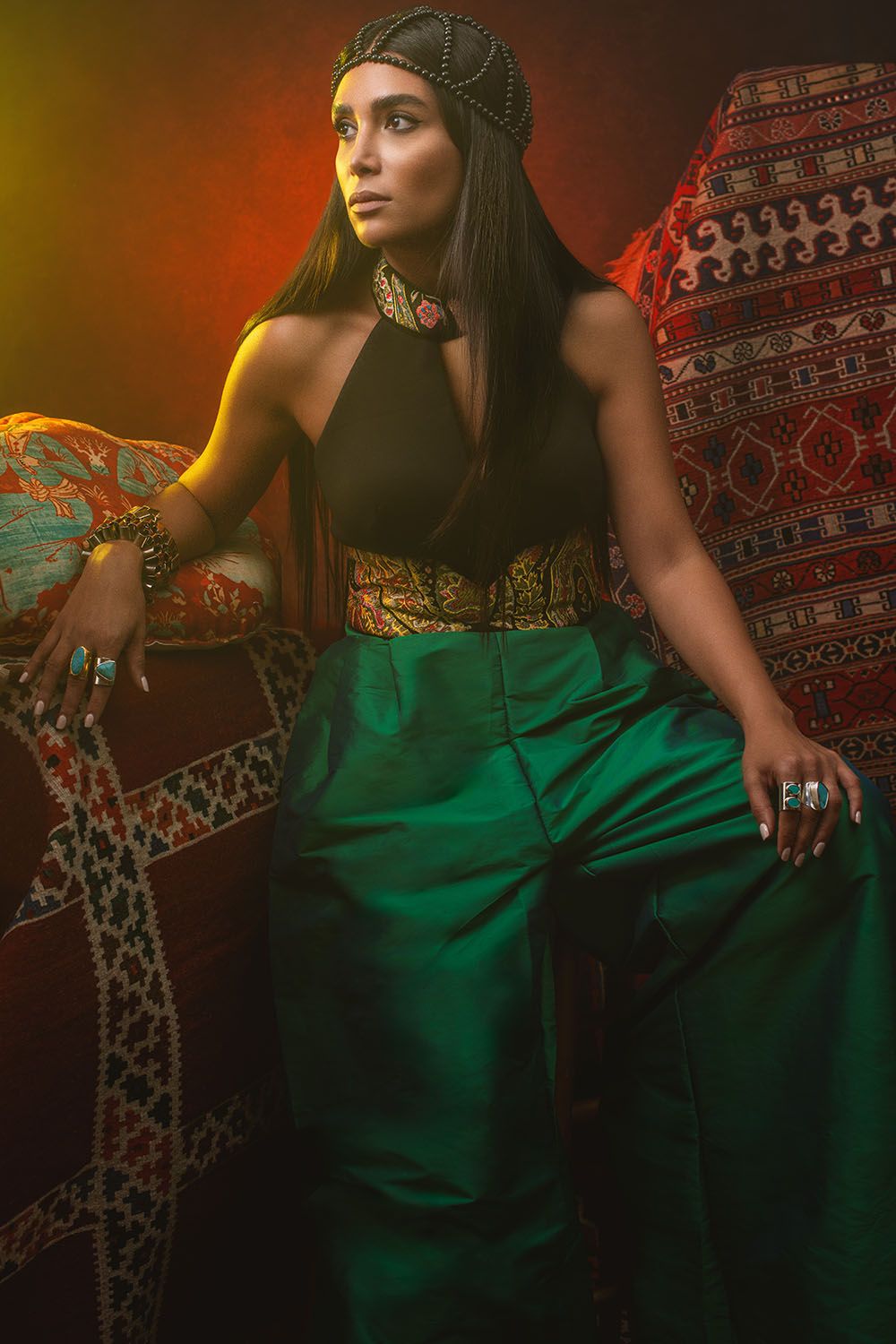 Woman in green satin pants, black top, gold belt, and jeweled headpiece poses regally against colorful patterned textiles.