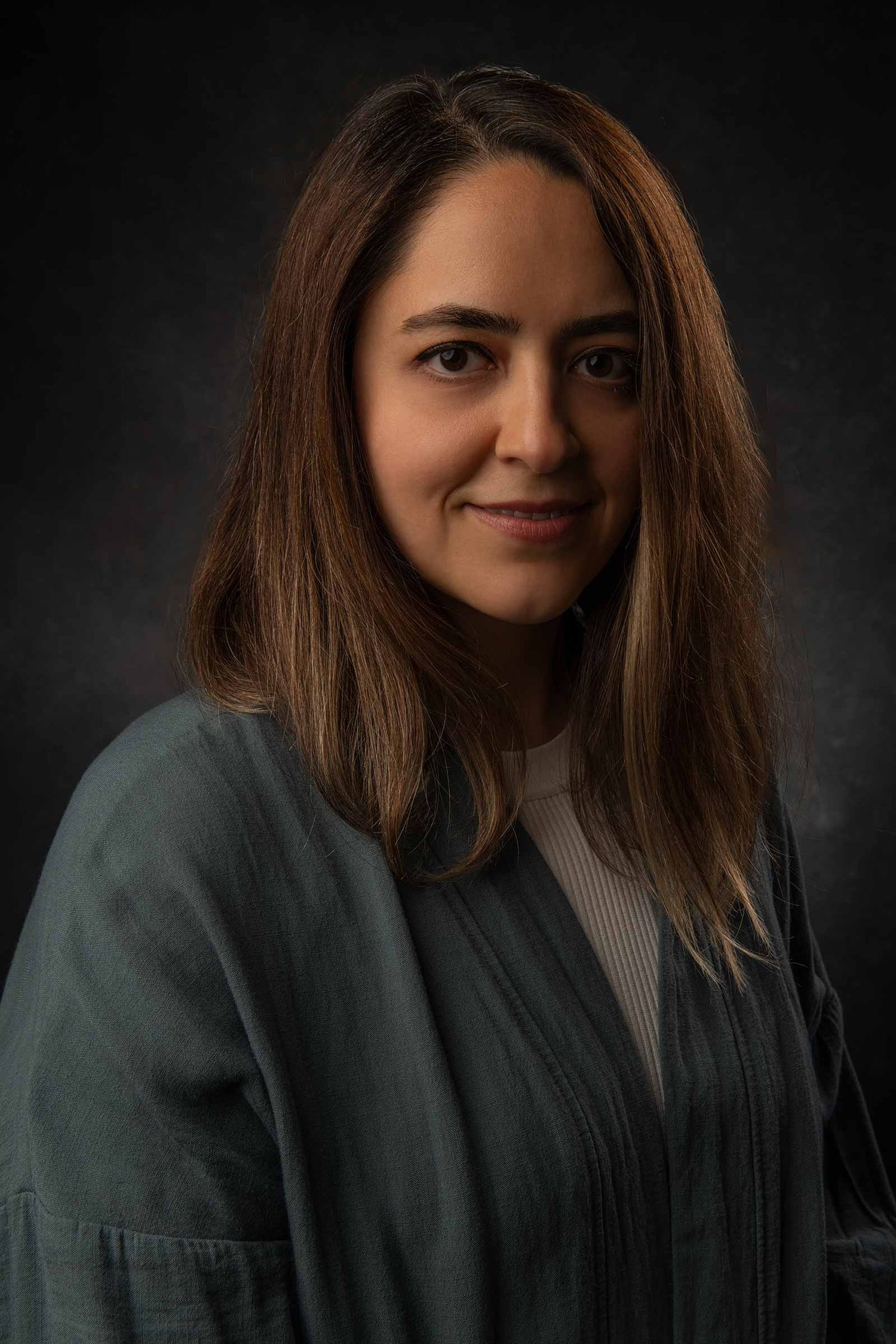 Professional portrait photo against dark background with person wearing sage green jacket and white top.