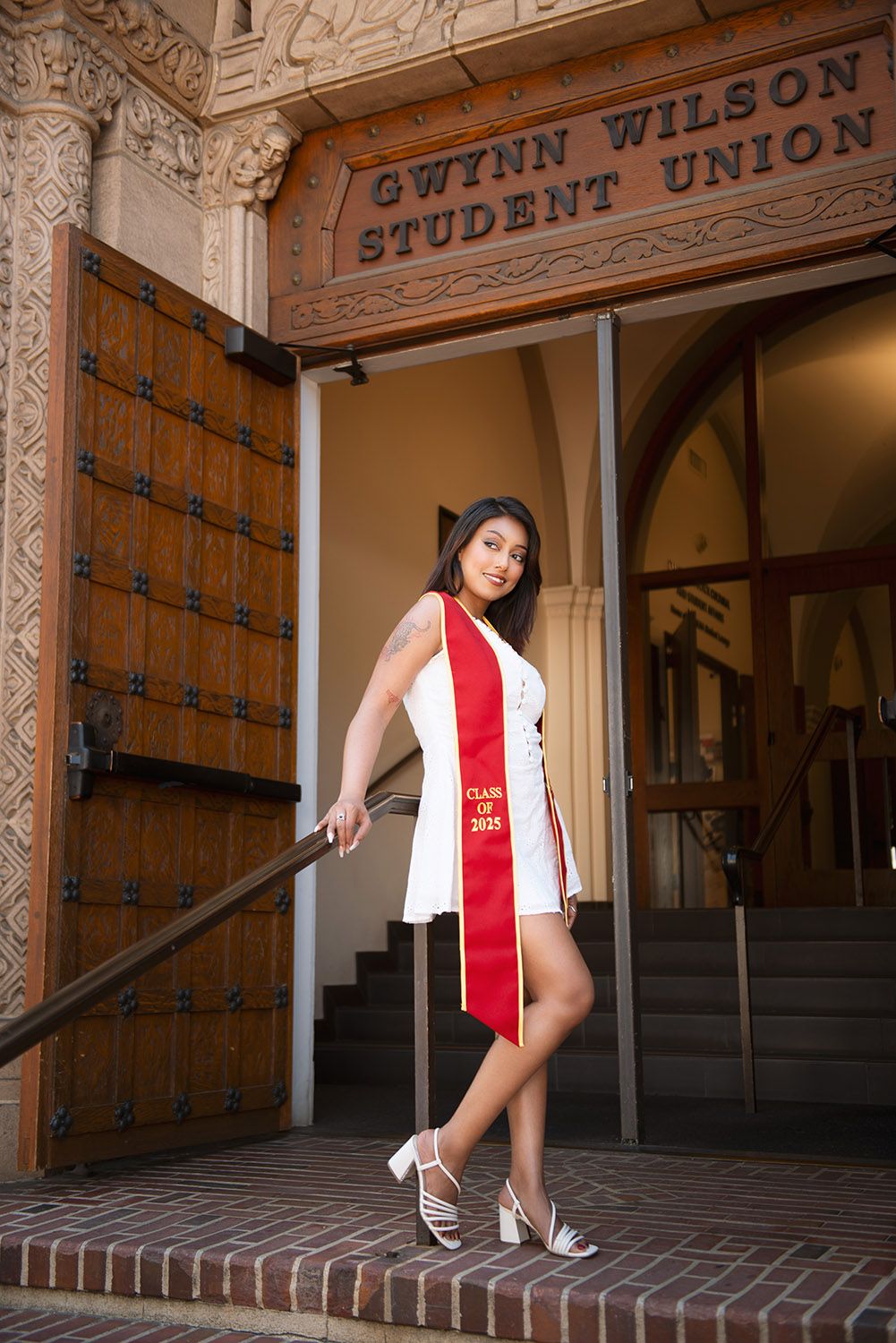 A graduate poses in a white dress and red Class of 2025 sash at Gwynn Wilson Student Union entrance.