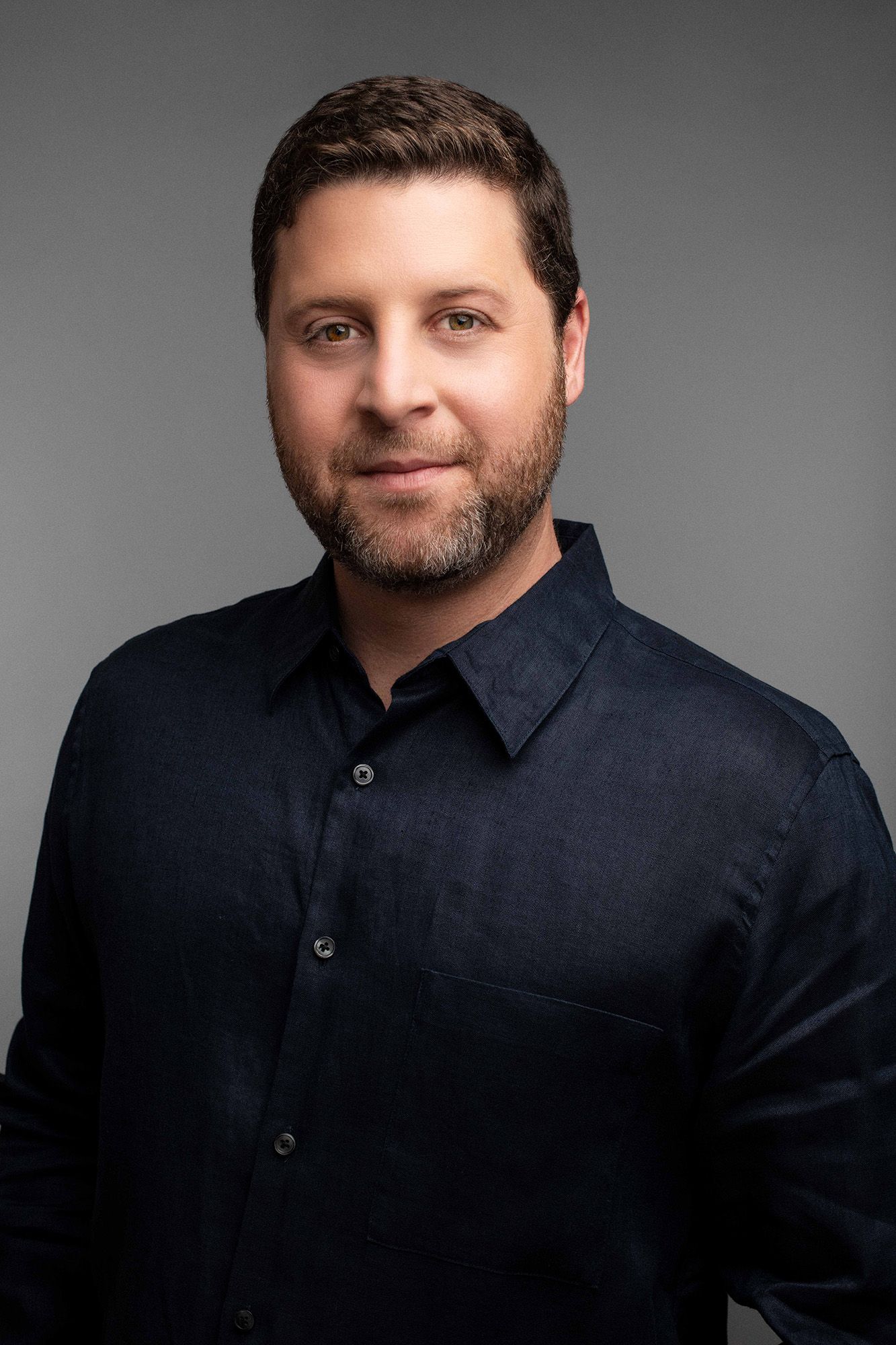 Professional headshot of a smiling man with dark hair and beard wearing a black button-up shirt against gray background.