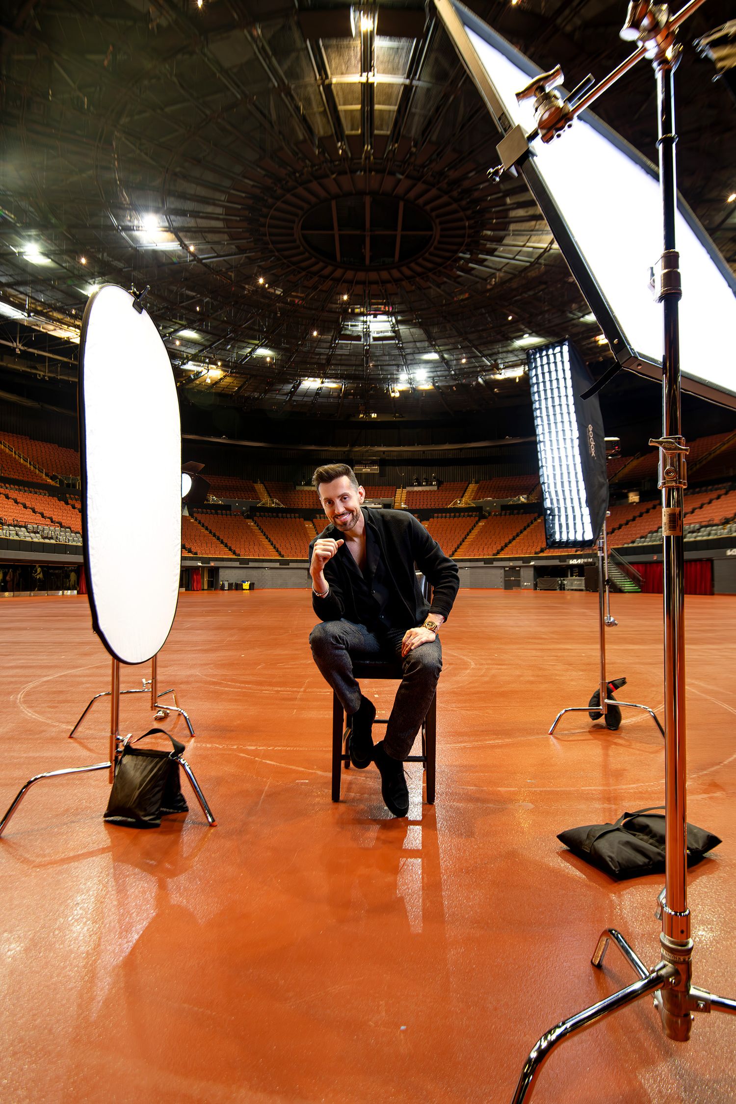 Behind the scenes portrait shoot on orange floor with lighting equipment and reflectors visible of comedian Max Amini in Kia forum.
