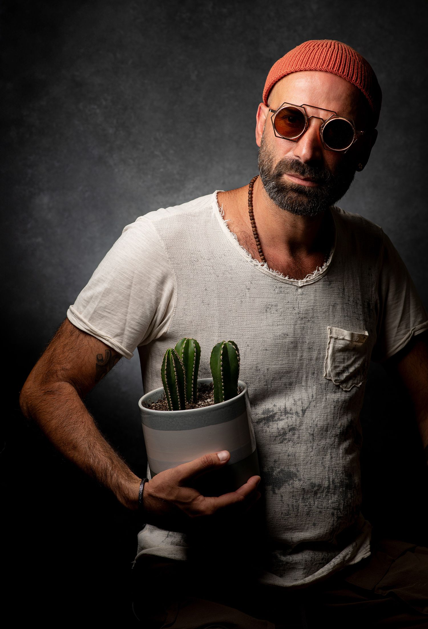 persian podcaster and comedian Farshid Manafi in gray t-shirt and orange beanie poses with houseplants in artistic studio shots.