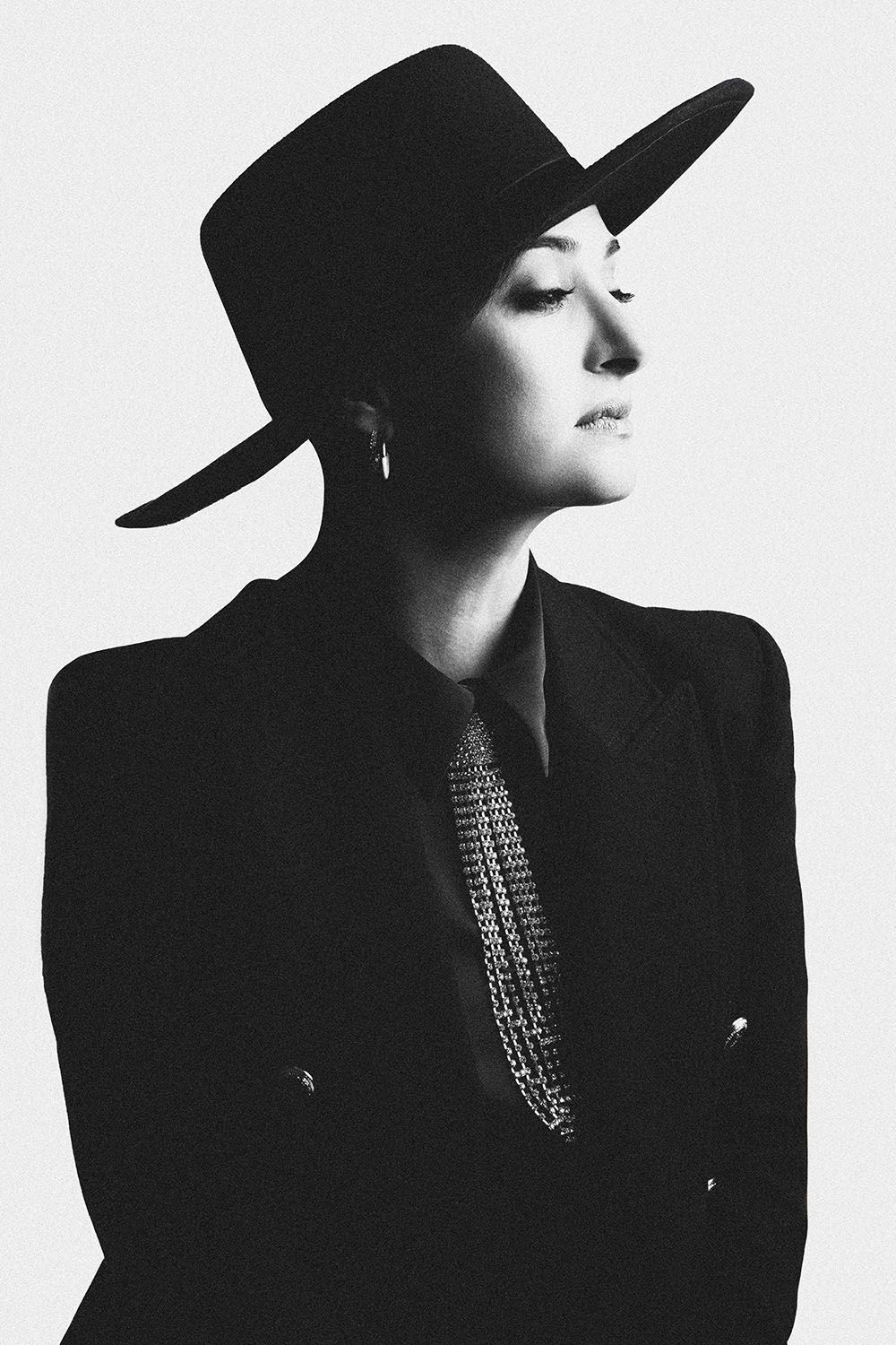 Black and white portrait of a woman in a wide-brim hat, black blazer, and layered chain necklace.