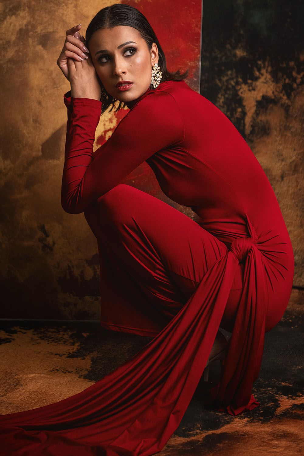 Woman in elegant red jumpsuit posing gracefully against a textured gold and dark background, wearing statement earrings.