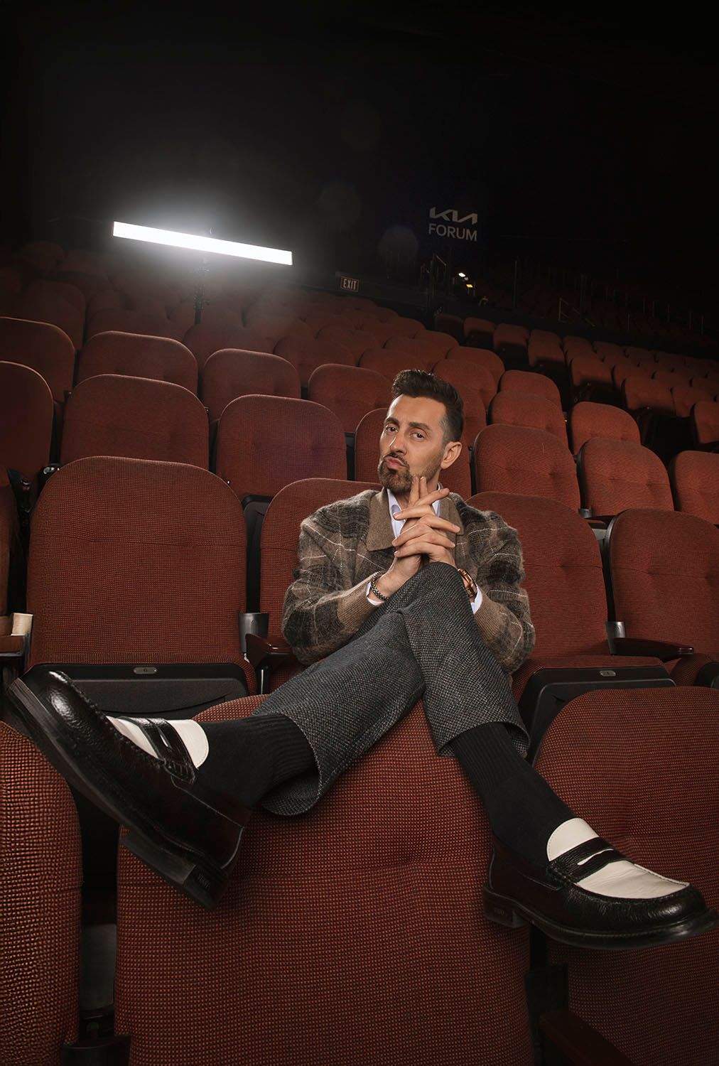 Man in plaid blazer sitting alone in red theater seats at Kia Forum, legs crossed, thoughtful pose.