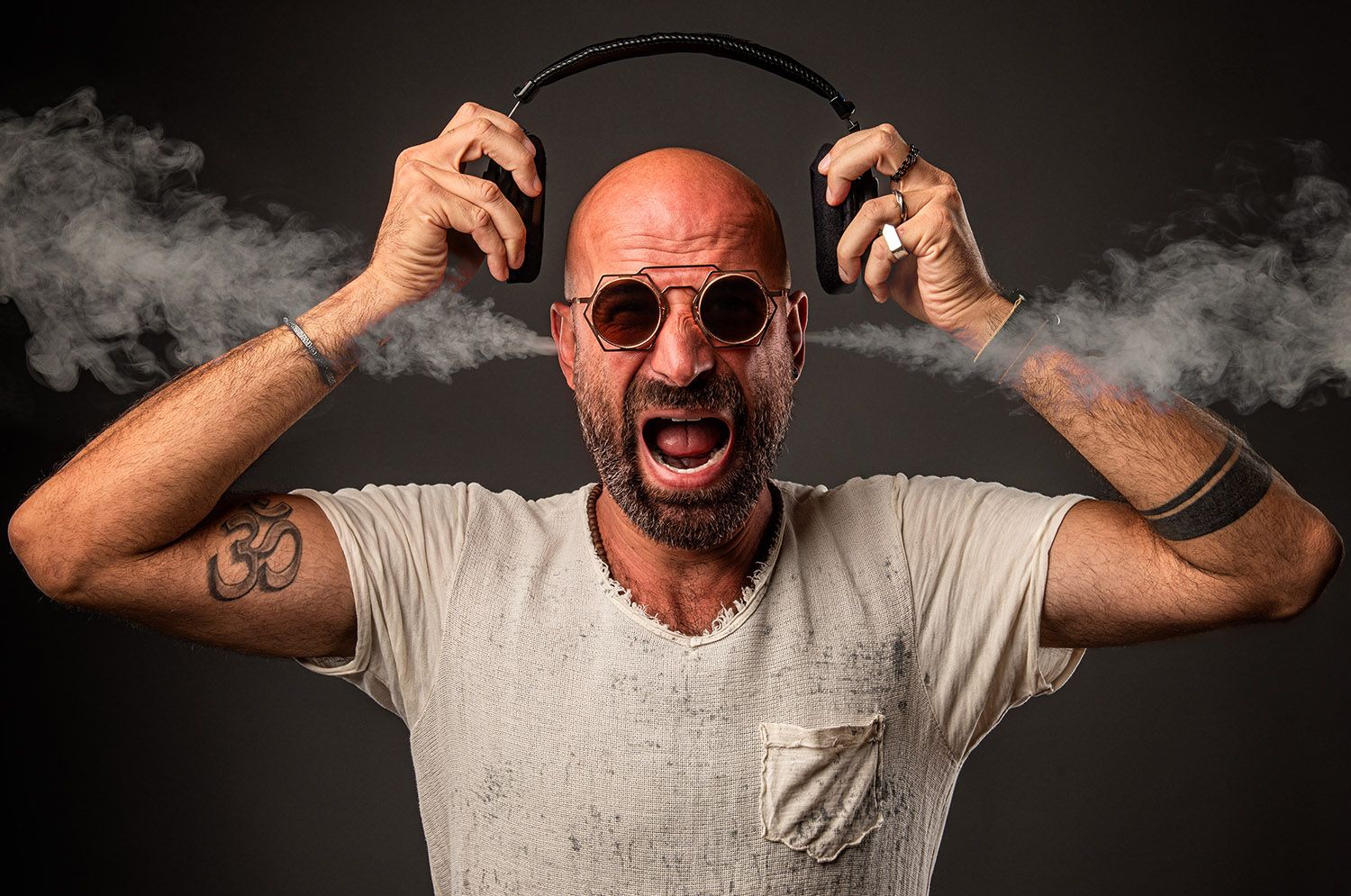 Bald man with beard and sunglasses screaming while holding headphones with smoke effects.
