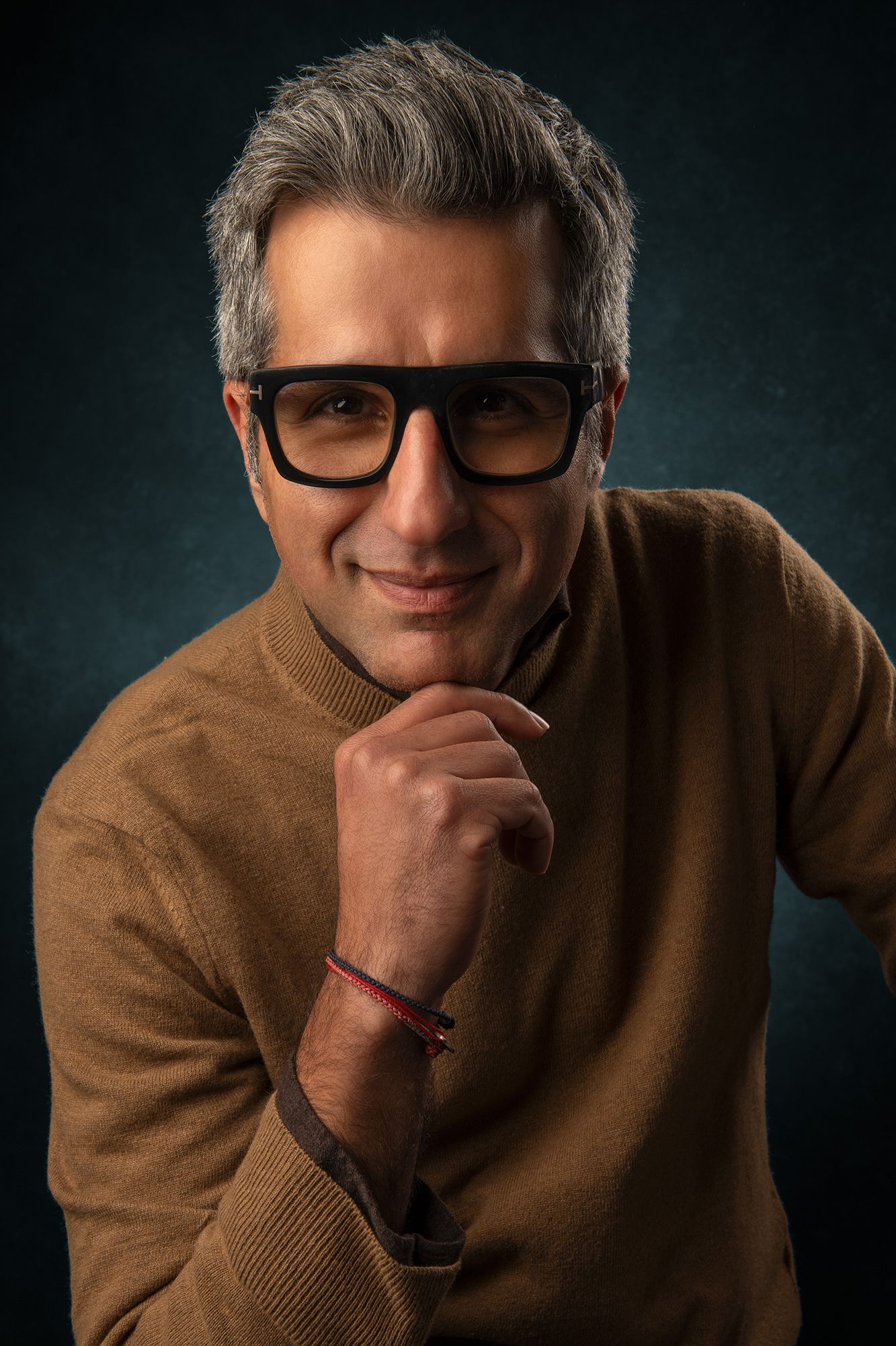 Studio portrait of person in brown sweater and glasses with hand on chin against dark backdrop taken in west Los Angeles.