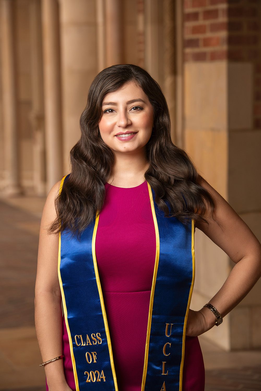 UCLA Class of 2024 graduate smiling in blue and gold sash over pink dress, posing in front of brick columns.