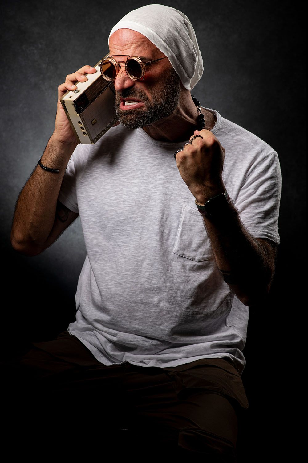 Bearded man in white tee and sunglasses holding a boombox radio to his ear with fist raised.