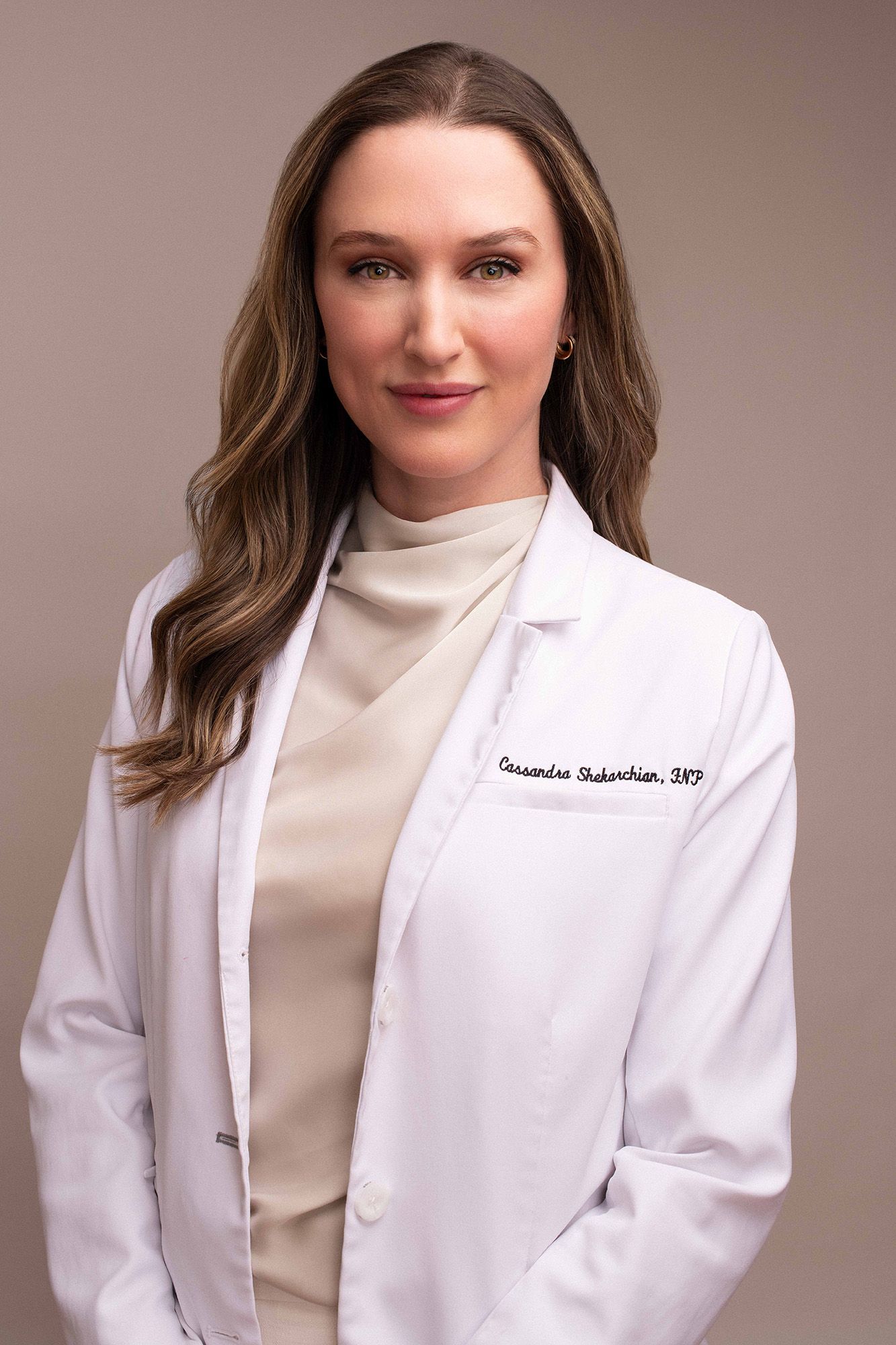 Female healthcare professional in white lab coat with name tag, posing confidently against neutral background.