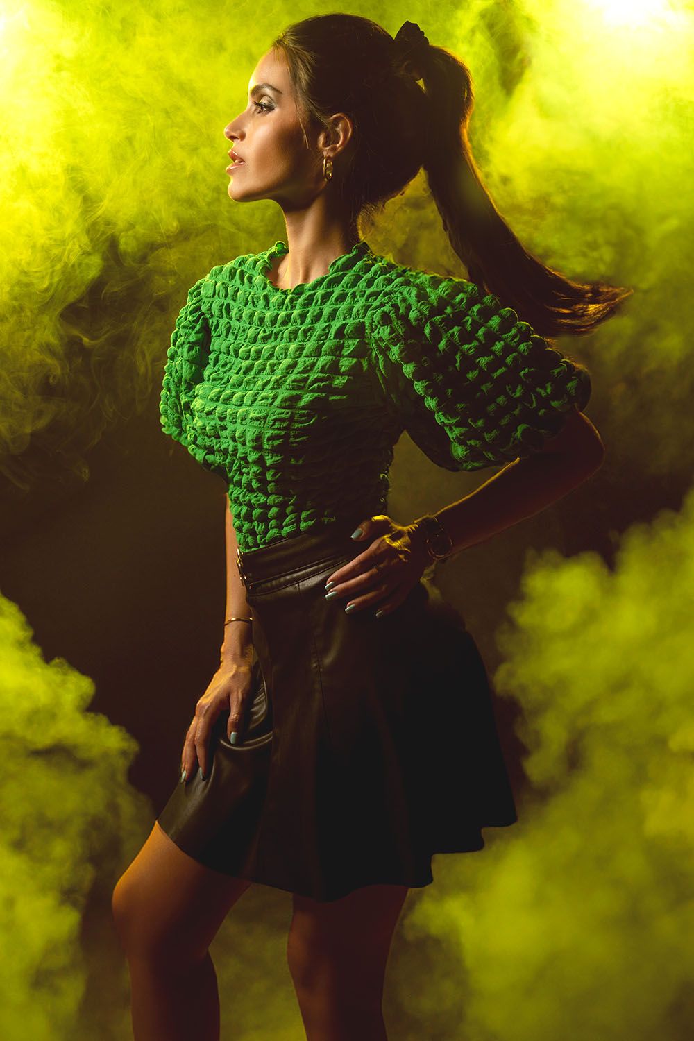 Woman in green textured top and black leather skirt posing dramatically in yellow-green smoke on dark background.