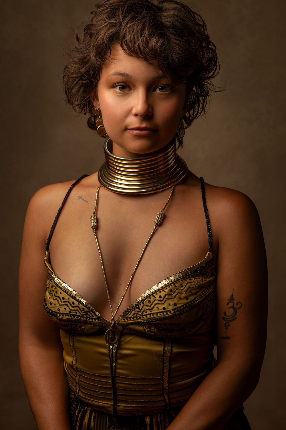 Woman in gold tribal outfit with brass neck rings and curly hair poses against dark background.