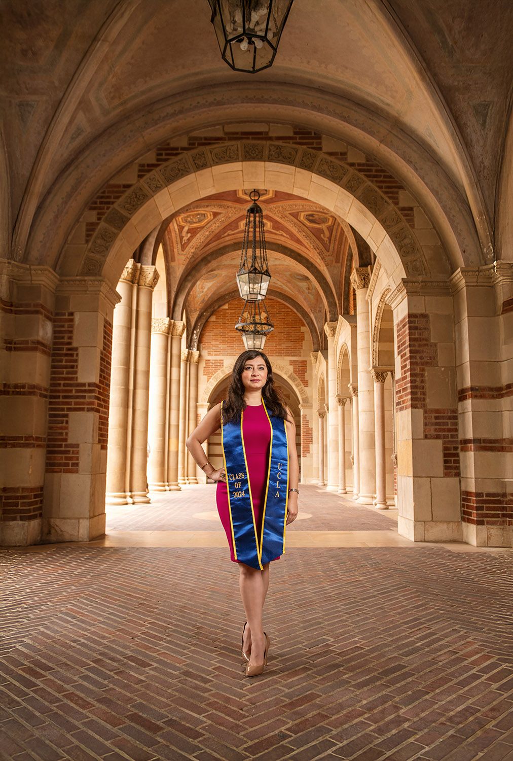 Graduate in cap stole poses under ornate arched colonnade of UCLA Royce Hall brick corridor.