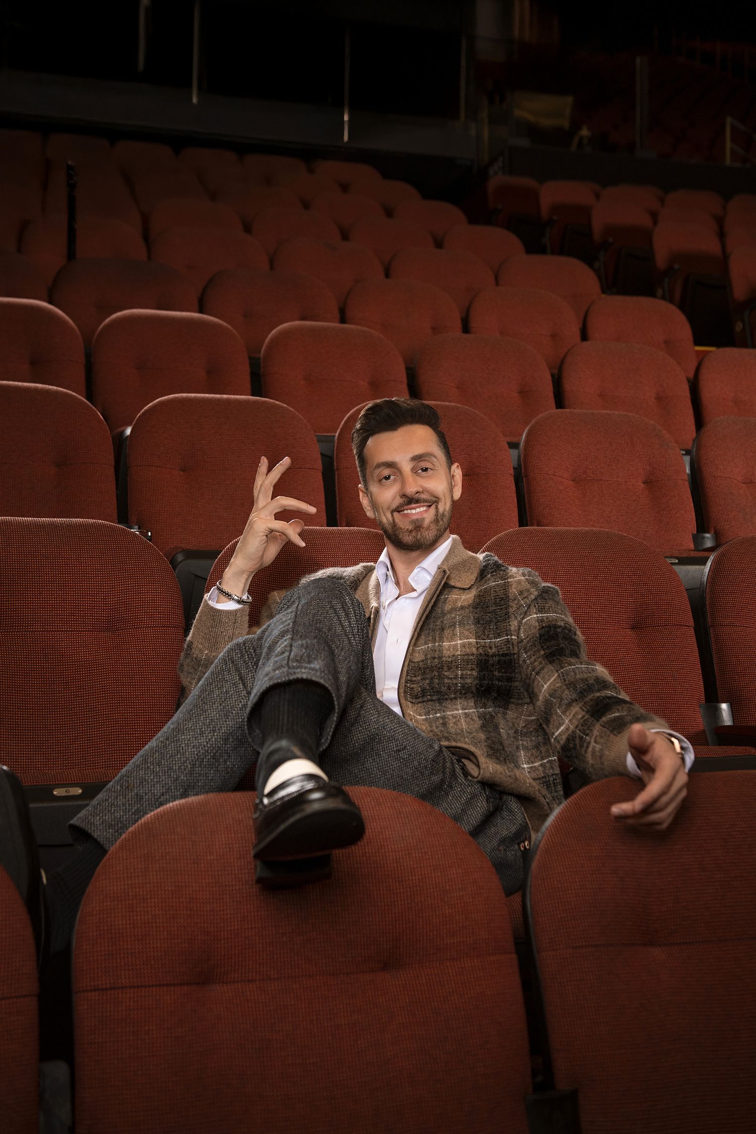 Casual portrait of persian comedian Max Amini lounging across theater seats in plaid jacket in Kia forum.