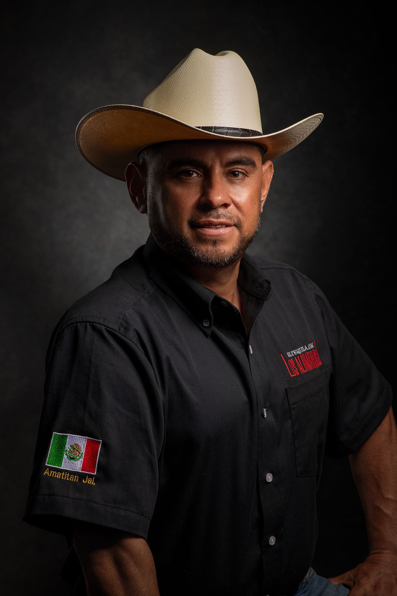 dramatic Portrait and headshot of person wearing cowboy hat and black shirt with Mexican flag patch against dark background.
