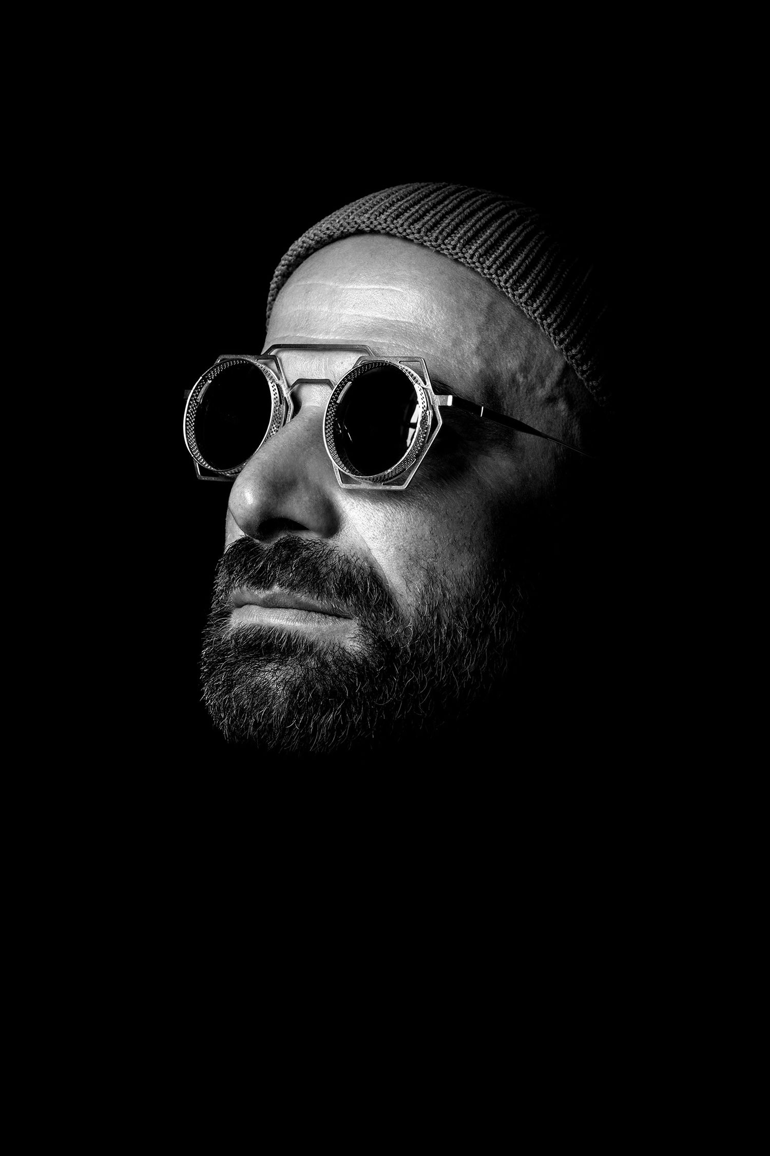 Dramatic black and white portraits of persian podcaster and comedian Farshid Manafi wearing beanie and round sunglasses.