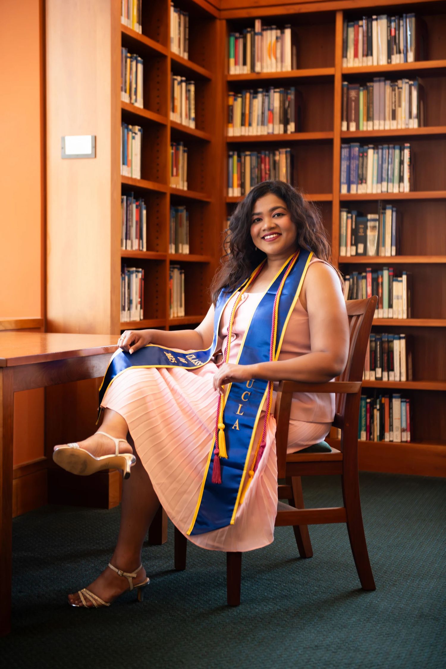 UCLA graduate in blue and gold stole sits smiling in library chair surrounded by wooden bookshelves.