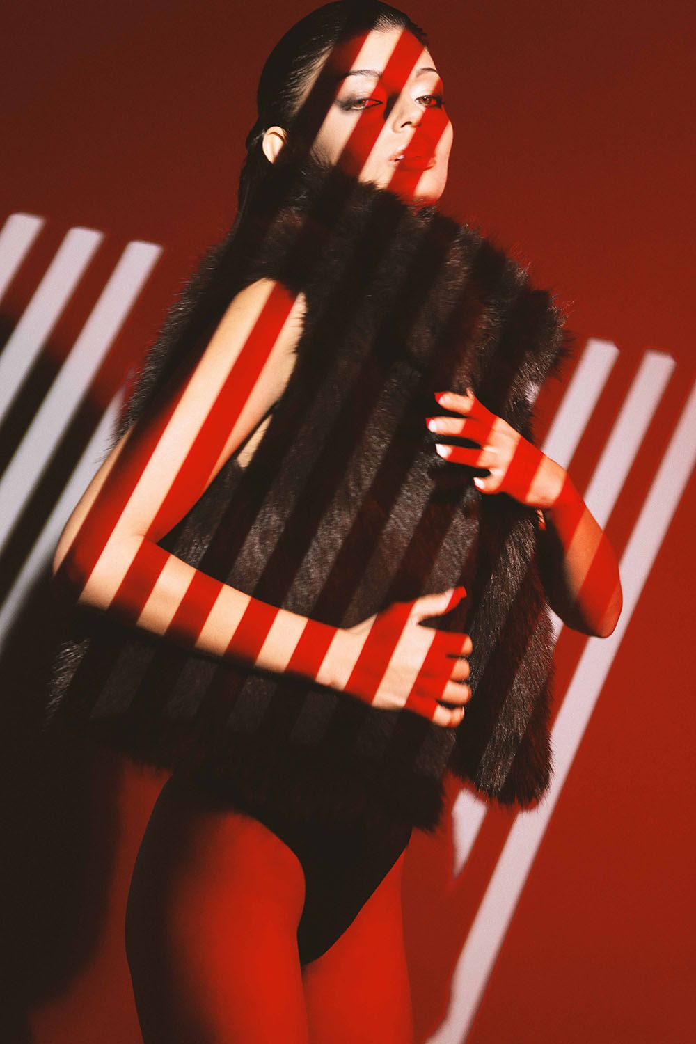 Woman in fur vest poses against red background with dramatic striped shadow light pattern creating bold visual contrast.