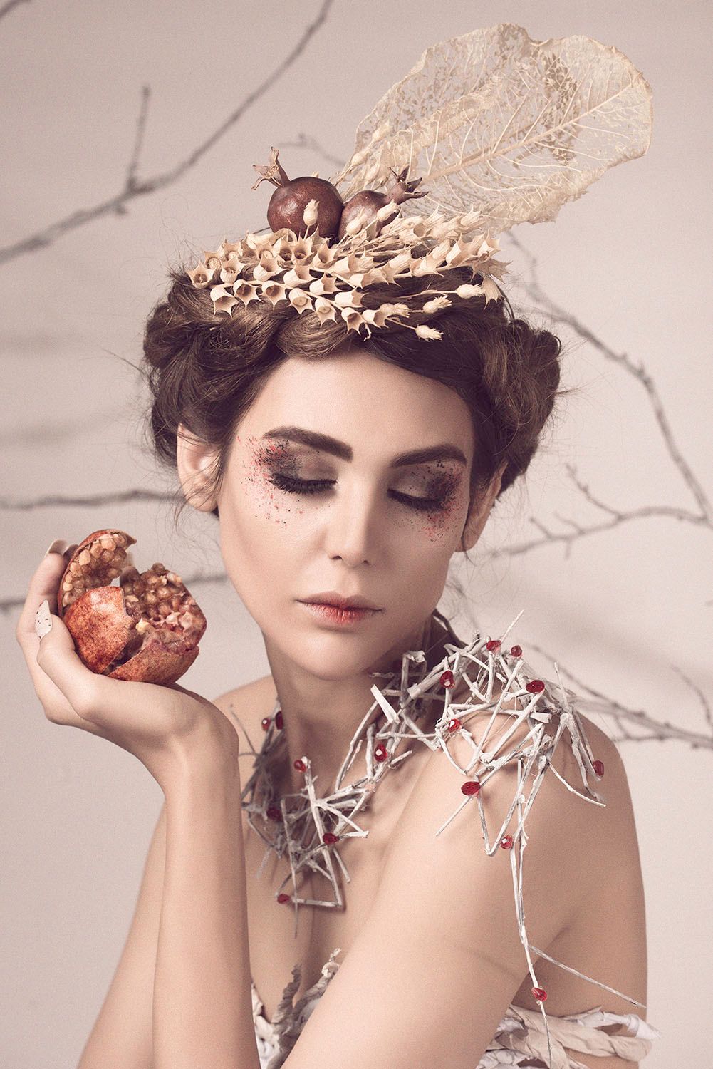Woman with artistic headdress of dried botanicals holds a pomegranate, wearing an avant-garde twig necklace in a fantasy portrait.