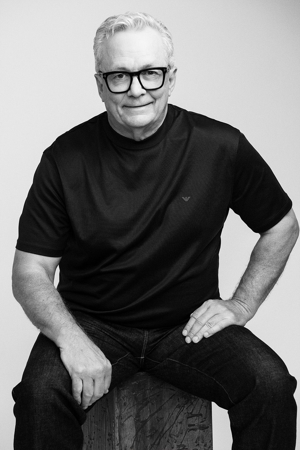 Black and white portrait of a senior man with gray hair, glasses, and black t-shirt sitting confidently.