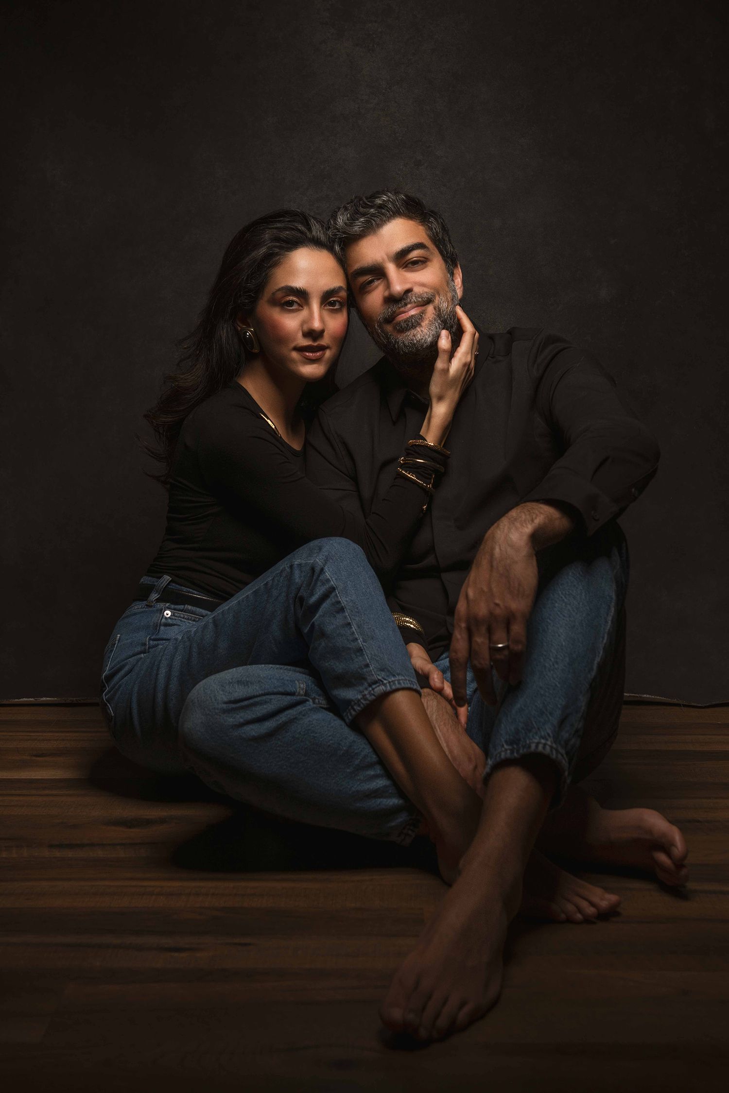 A couple in black outfits and jeans sit intimately together on a dark wooden floor in moody lighting.