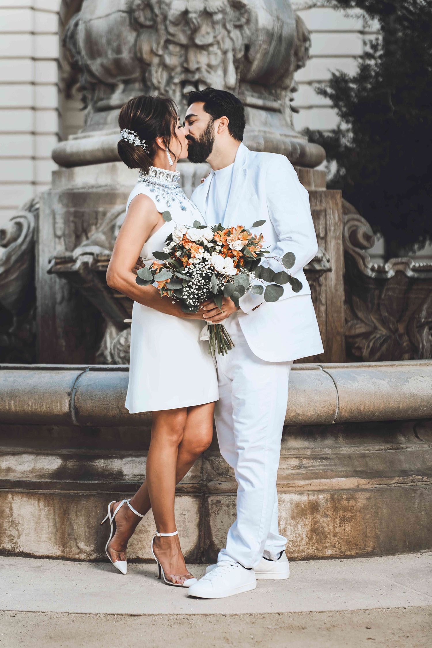 A romantic couple in all white outfits share a kiss by ornate stone architecture while holding a beautiful floral bouquet.