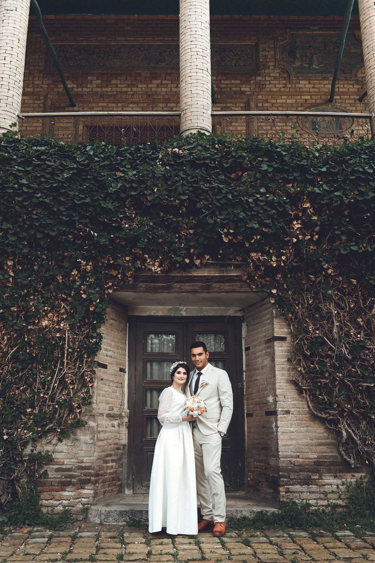 A couple in white lab coats stands together against a brick building with climbing ivy.