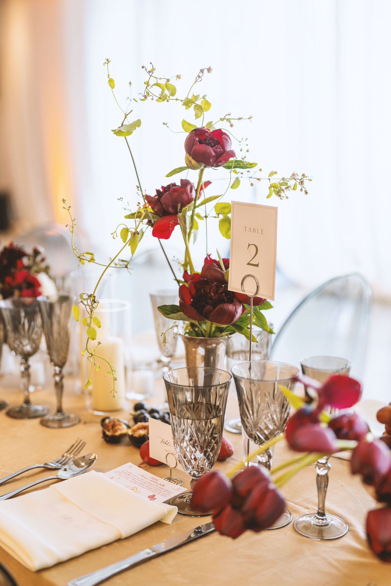 Elegant table setting with burgundy roses in crystal vases and red apples as decor elements on white tablecloth.