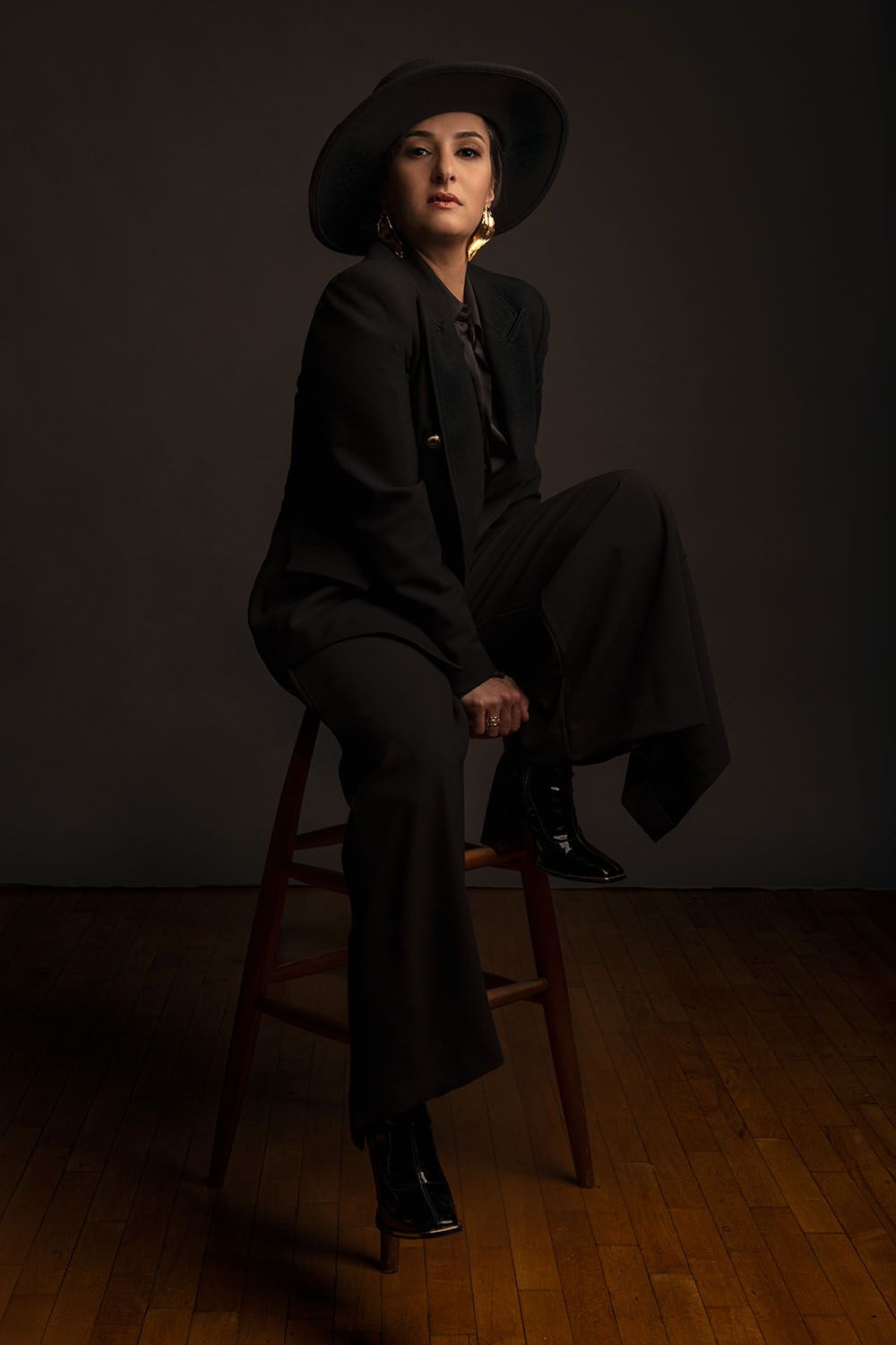 Woman in black suit and wide-brim hat posing on wooden stool against dark background.