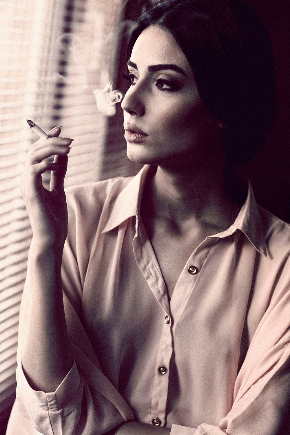 Elegant woman in pink blouse smoking cigarette by window blinds in moody black and white portrait.