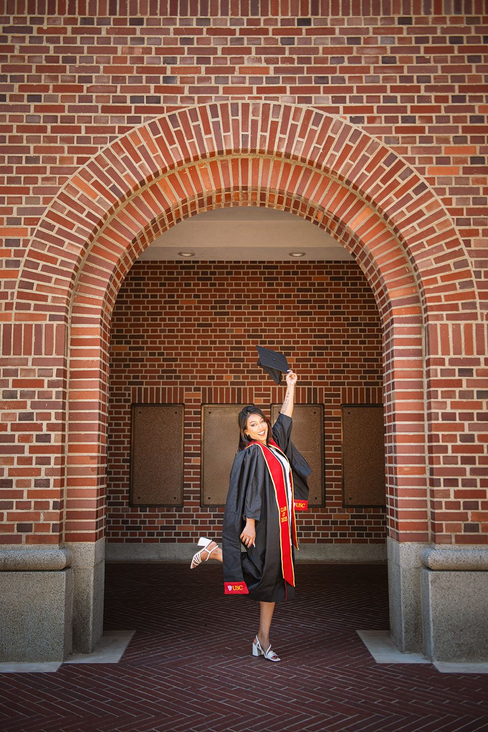 Graduate in black cap and gown celebrates under a red brick archway, raising cap triumphantly.