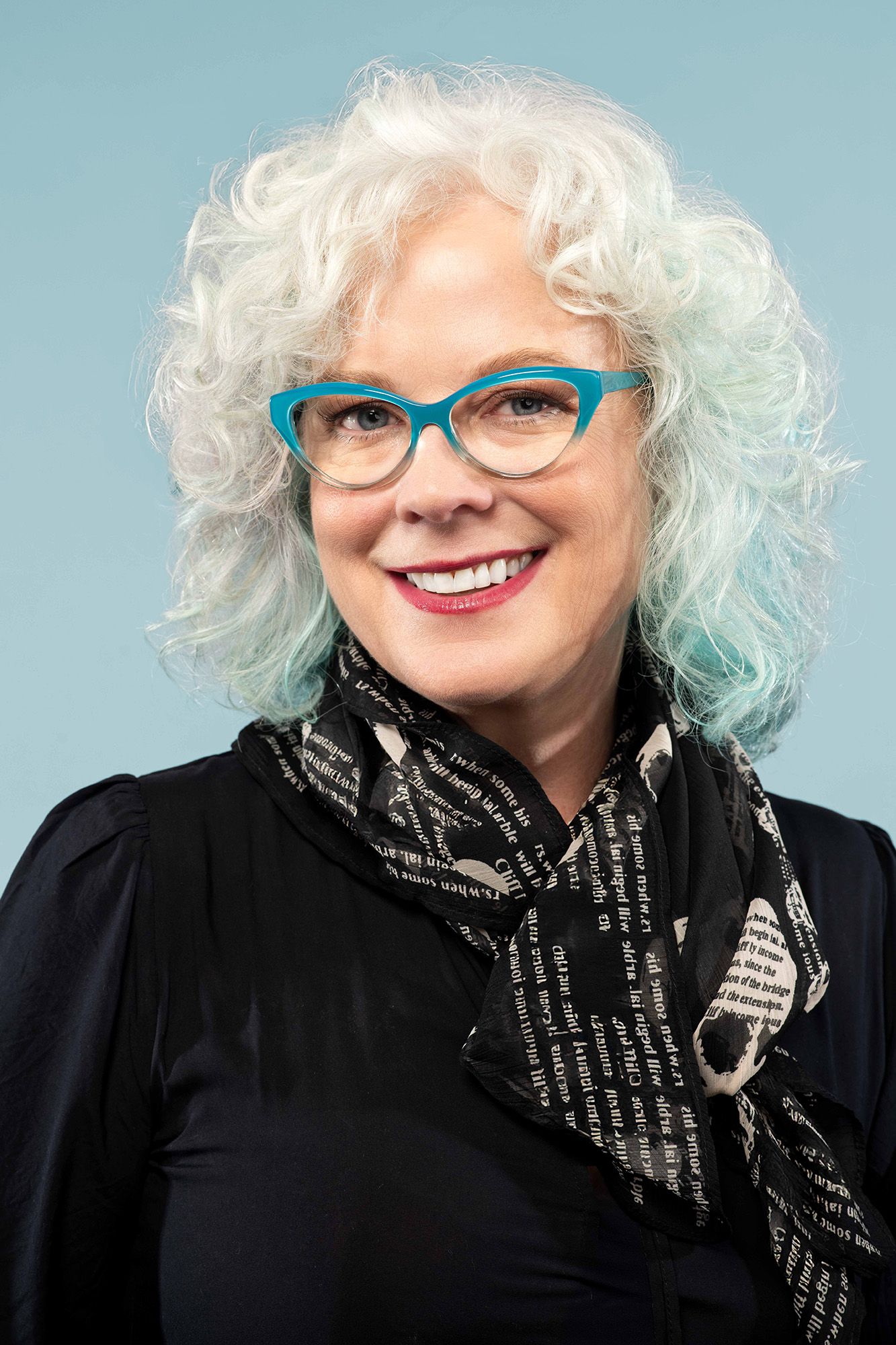 Smiling woman with curly white hair, teal cat-eye glasses, black blouse, and printed scarf on blue background.