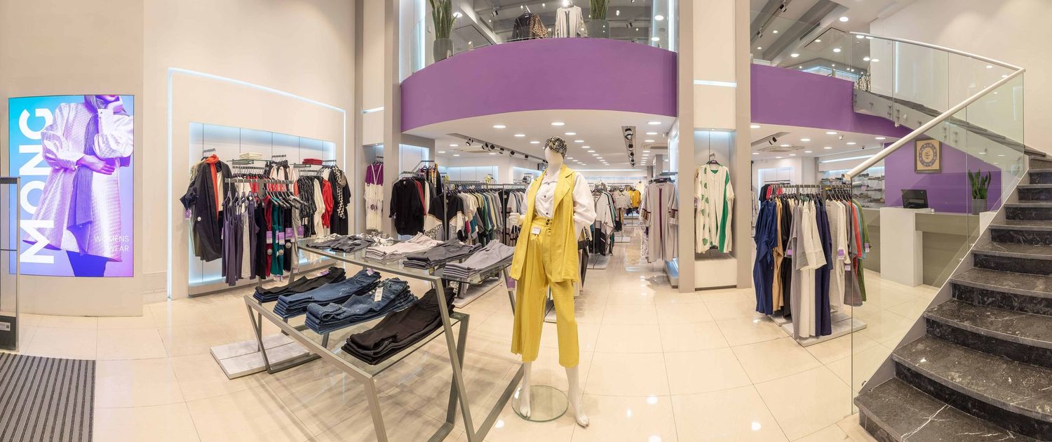 Modern retail clothing store interior with mannequins, hanging racks and a curved staircase under purple lighting fixtures.