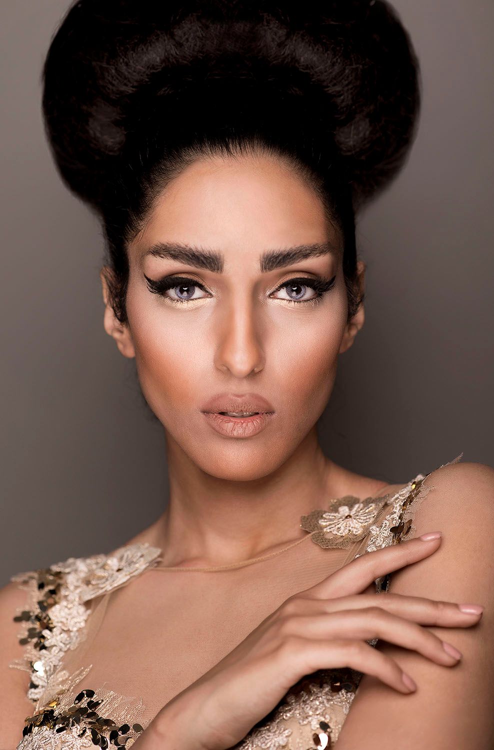 Elegant woman with bold winged eyeliner and voluminous updo hairstyle wearing gold lace dress in studio portrait.