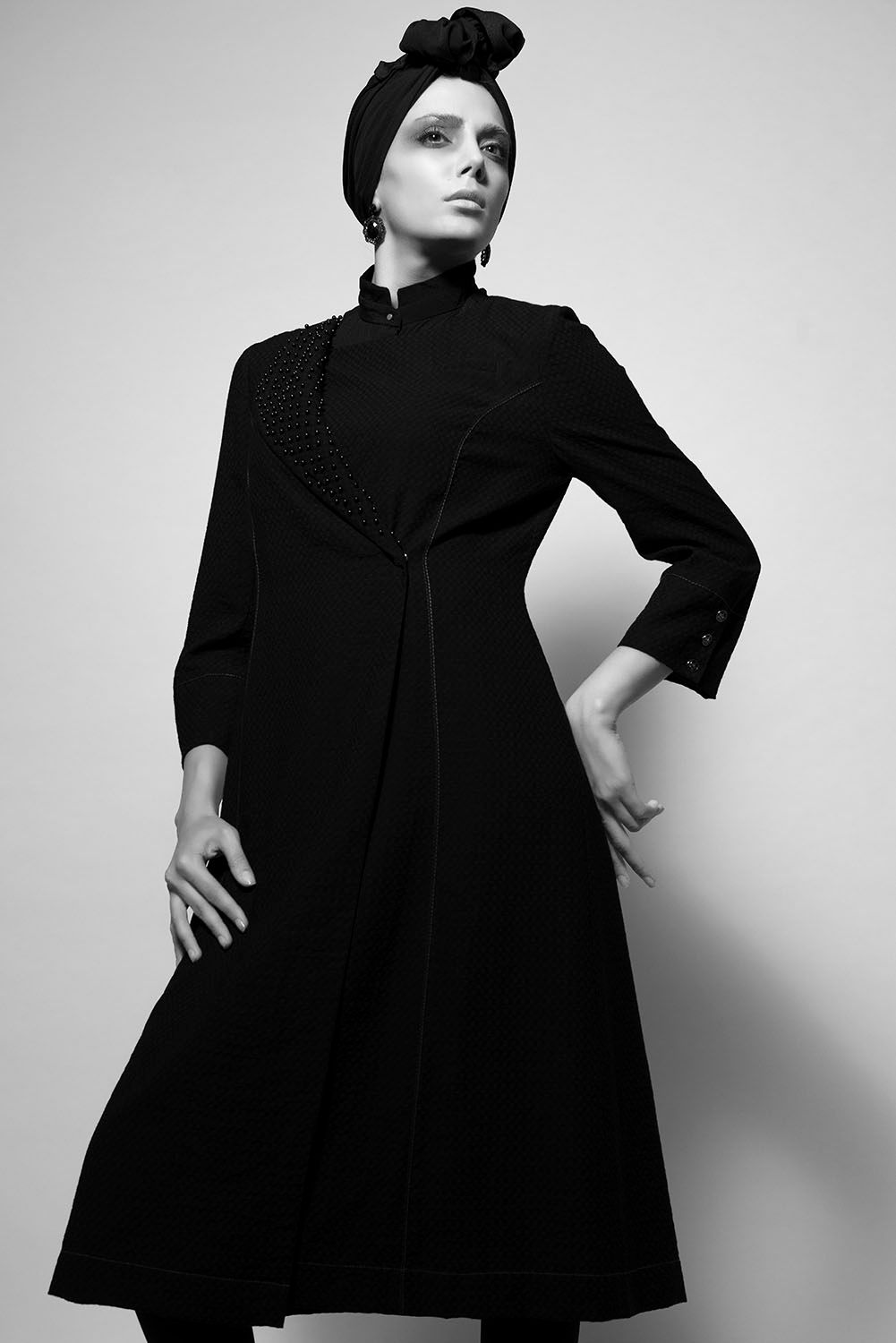 Black and white photo of a woman in a black coat and turban headwrap, posing with hands on hips.