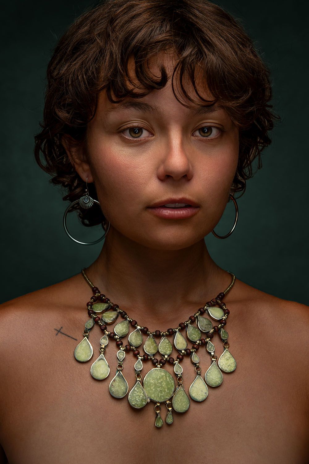 Young woman with curly hair wearing a green stone necklace and hoop earrings against a dark background.