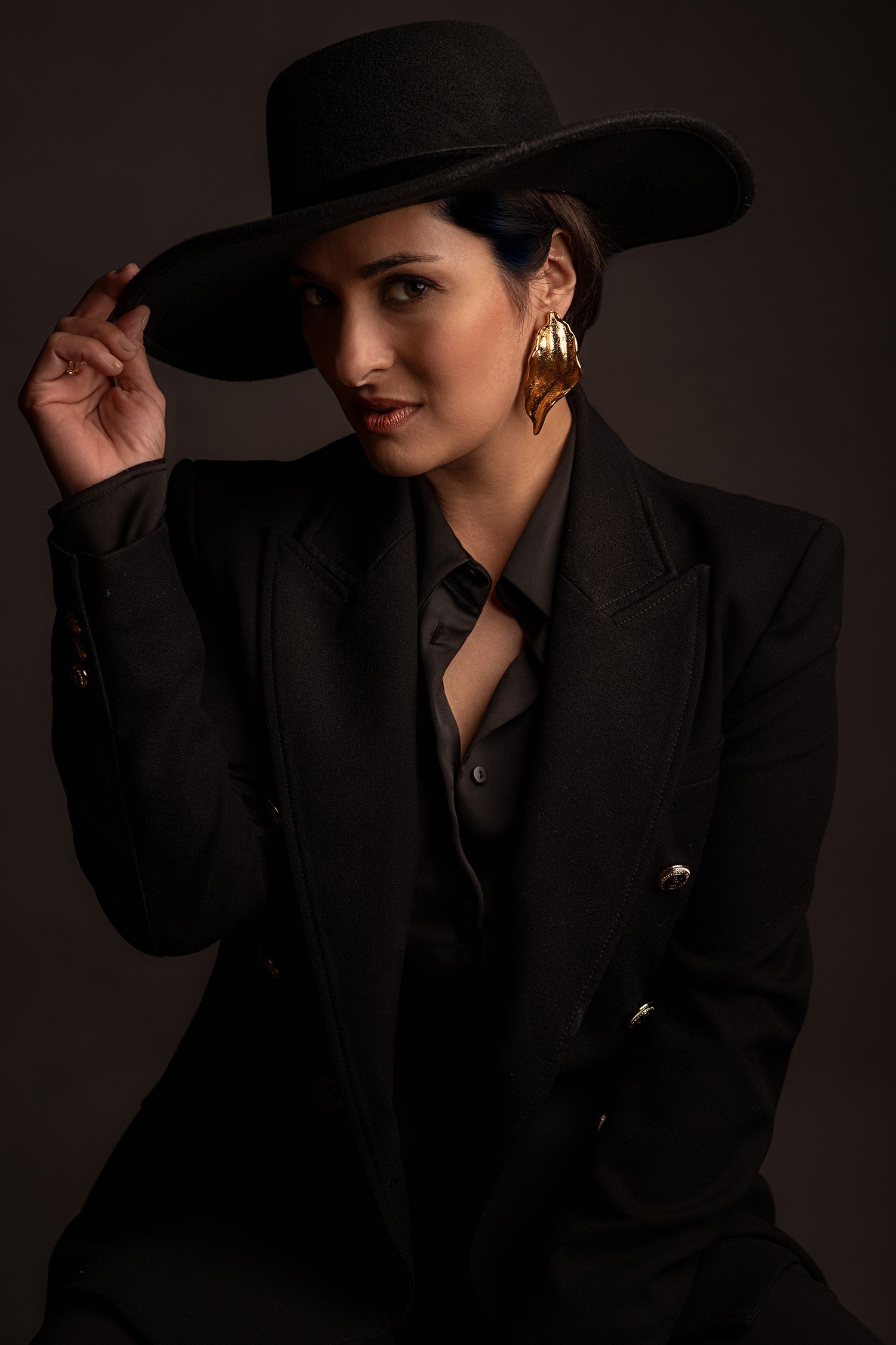 Moody fashion portraits of persian pop singer Rana Mansour in black blazer and wide-brim hat against dark background.