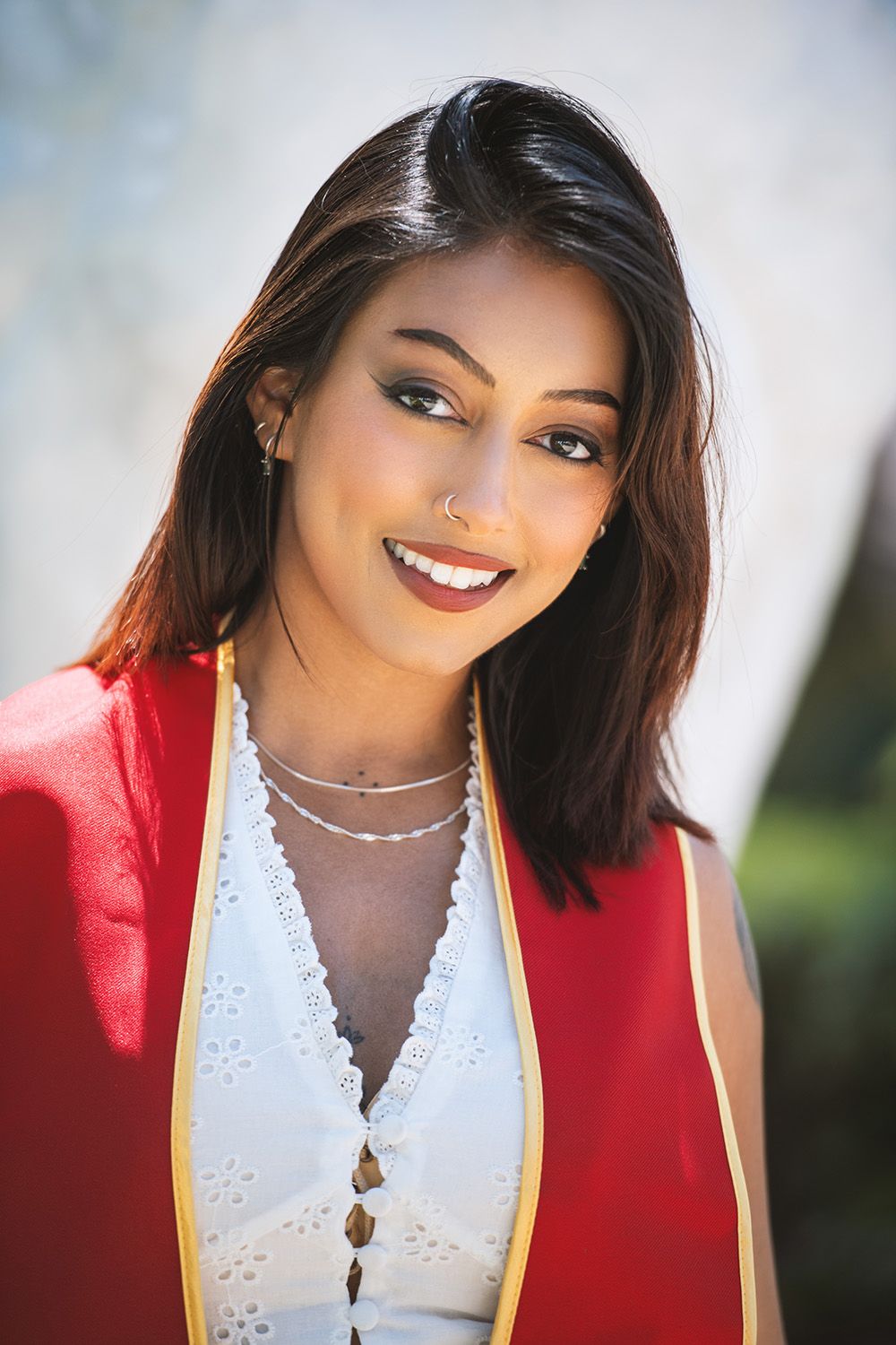 Young woman in red graduation gown smiling outdoors, wearing white dress and silver necklace.