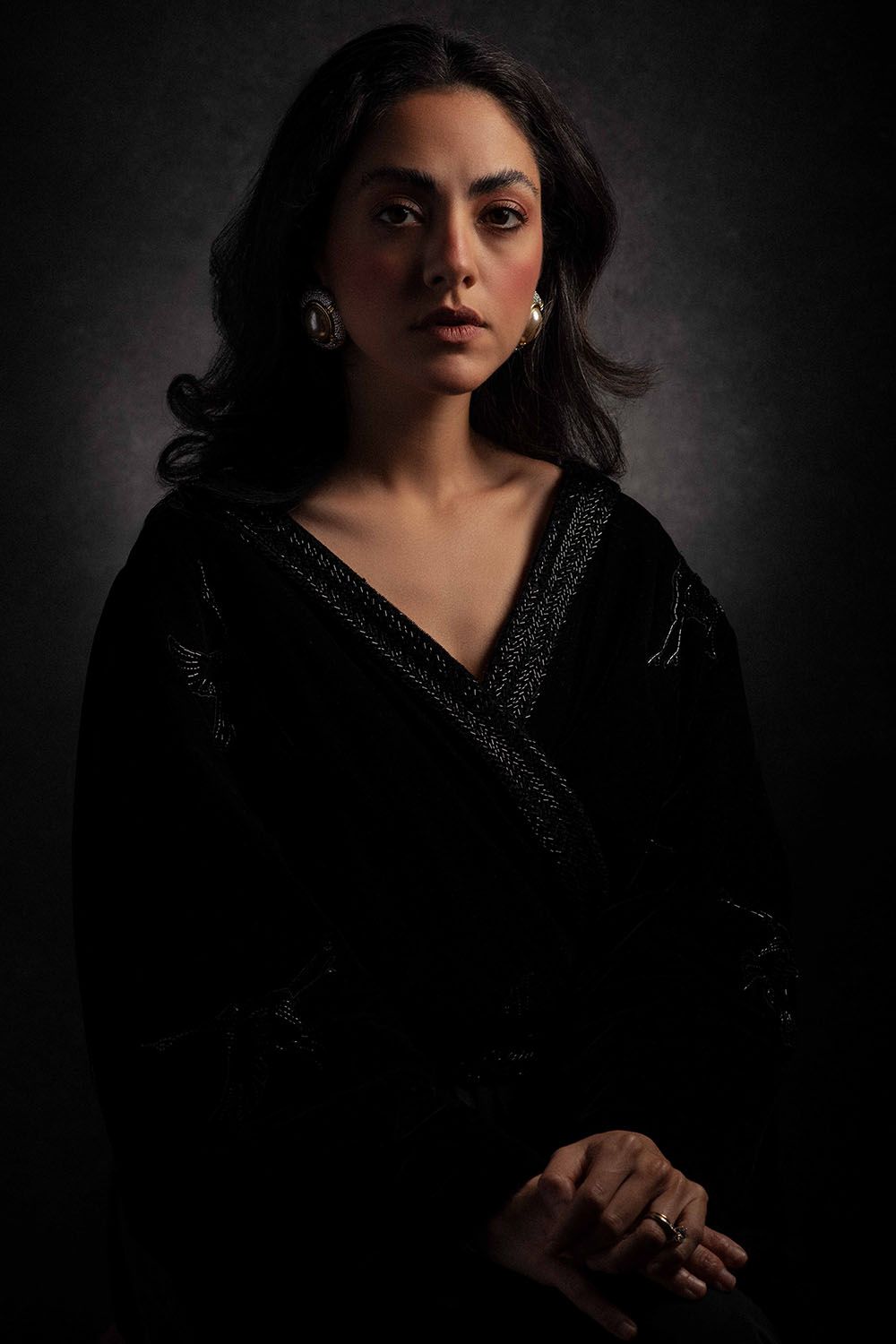 Dark-haired woman in black velvet outfit with silver earrings posing against gray background.
