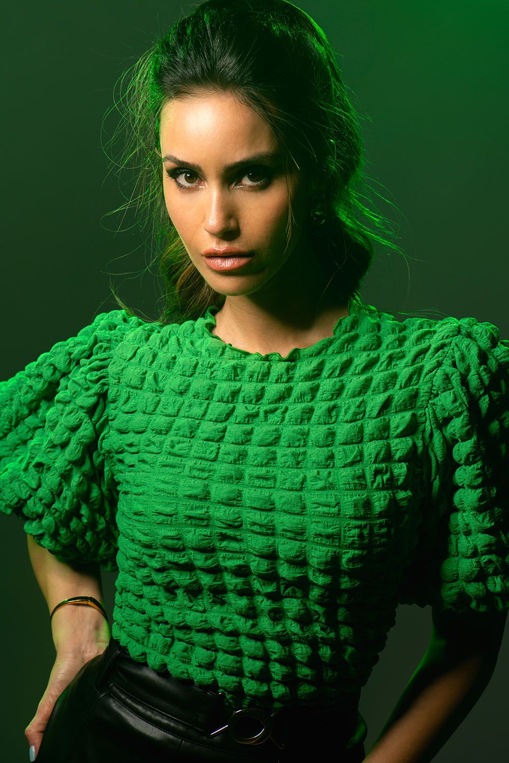 Woman in green textured puff-sleeve top posing confidently against a dark green background with dramatic lighting.