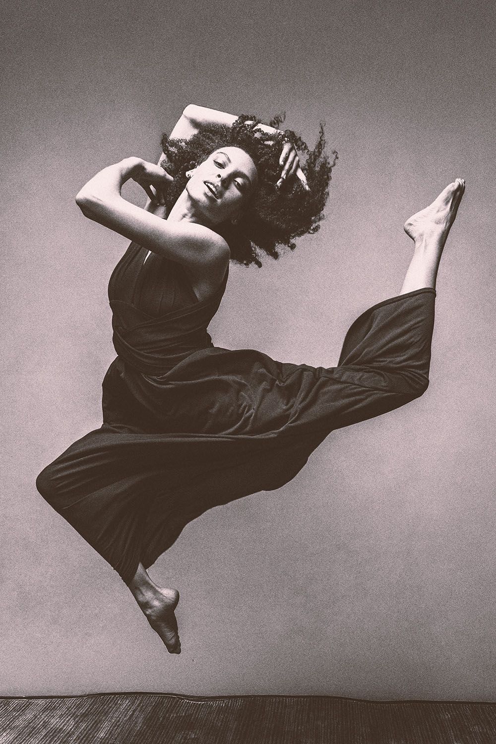 Black and white photo of a dancer mid-leap in a dark jumpsuit, arms raised and legs split against a gray background.