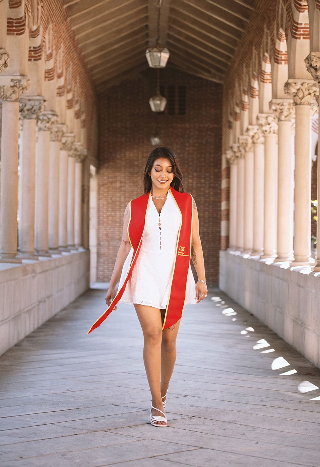 Graduate walks through elegant columned archway wearing white dress and red graduation sash at university.